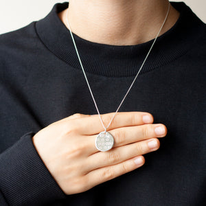PARAGON IV SMALL ROUNDISH SILVER PENDANT WITH SQUARE PATTERN ON CHAIN BY MARGO ORLOVIK ON A MODEL'S HAND